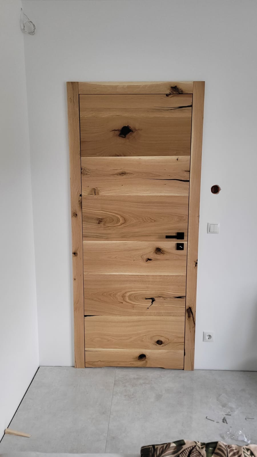 Dębowa stolarka drzwiowa z widocznymi sękami i czarnym klamką na tle białej ściany i szarej podłogi.