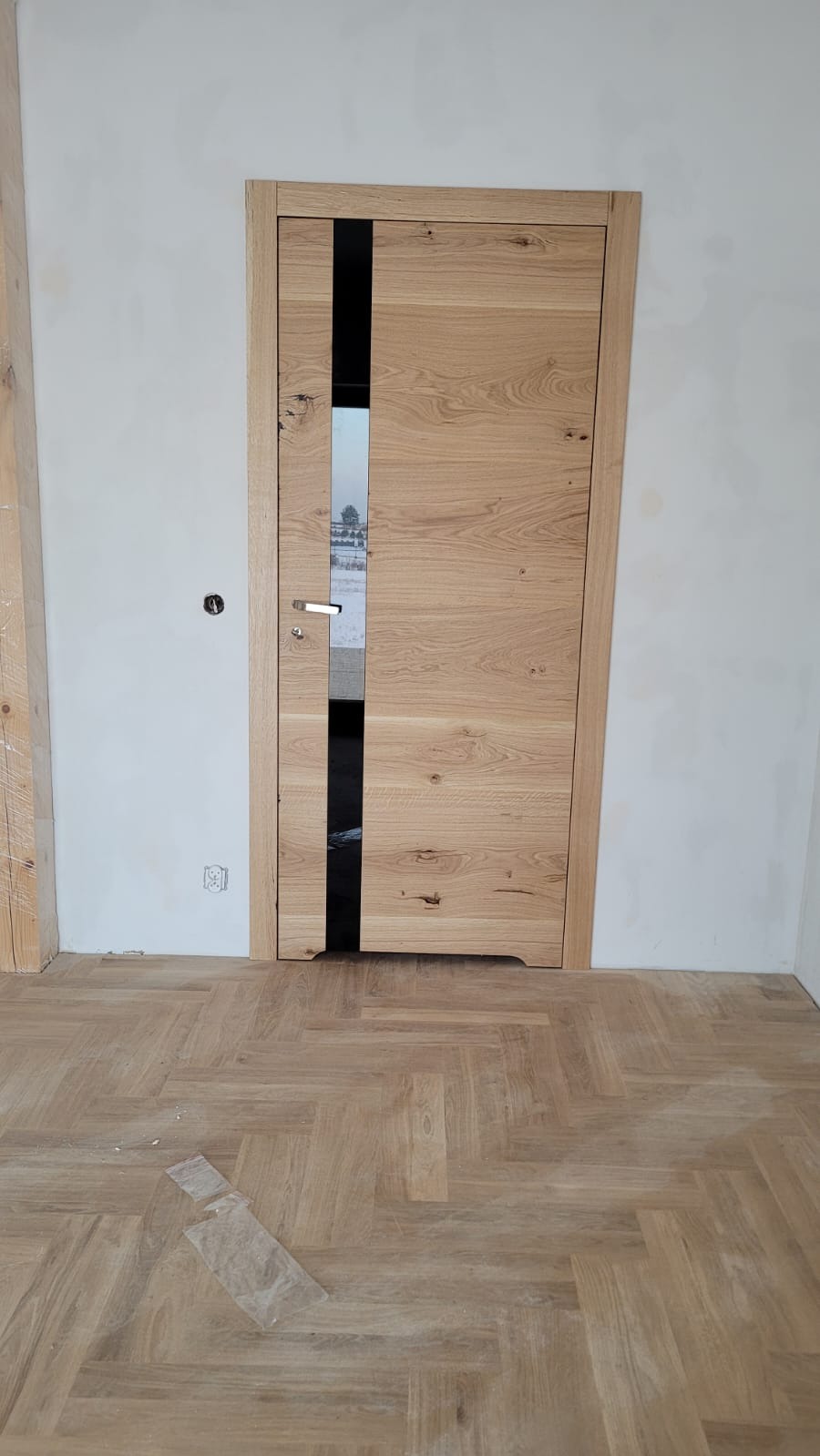 Otwarta, drewniana drzwi wewnętrzne z pionowym, czarnym przeszkleniem, zamontowana w surowym wnętrzu z parkietem w jodełkę, widoczne ślady prac wykończeniowych.