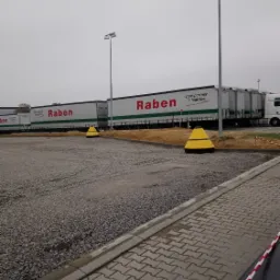 Flota ciężar&oacute;wek firmy Raben zaparkowana na placu wyłożonym szarą kostką brukową i żwirem, ograniczona ż&oacute;łtymi pachołkami i biało-czerwoną taśmą.