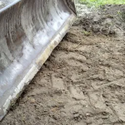 Metalowa łyżka koparki zagłębia się w błotnistą ziemię, widoczne ślady opon i świeża trawa w tle.