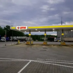 Stacja paliw Lotos z dystrybutorami, szarym brukowanym placem i zaparkowanymi samochodami ciężarowymi w tle pod pochmurnym niebem.
