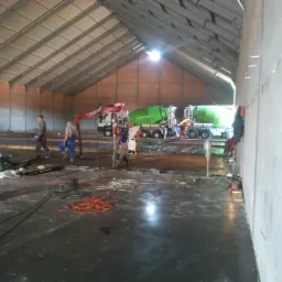 Wylewka posadzki przemysłowej w hali: pracownicy, betonowozy, rozłożone zbrojenie, przygotowanie do zalewania. Widok z boku, oświetlenie naturalne.