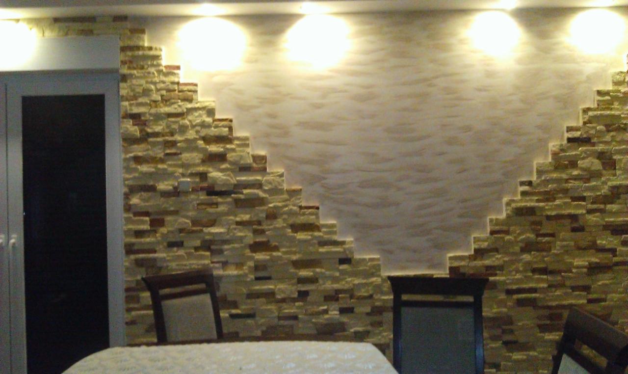 Dekoracyjna ściana z kamienia i tynku strukturalnego w ciepłych odcieniach, oświetlona punktowo, tworząca unikalny wzór w salonie. Widoczne krzesła i fragment stołu.