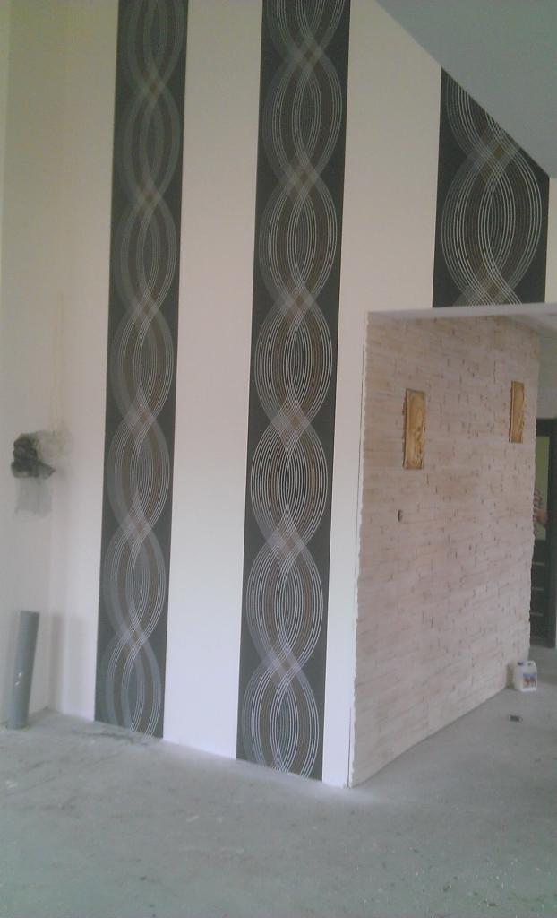 Wnętrze pokoju z dekoracyjną tapetą w geometryczne wzory i ścianą wyłożoną kamieniem dekoracyjnym, widoczne elementy wykończeniowe i surowy beton na podłodze.