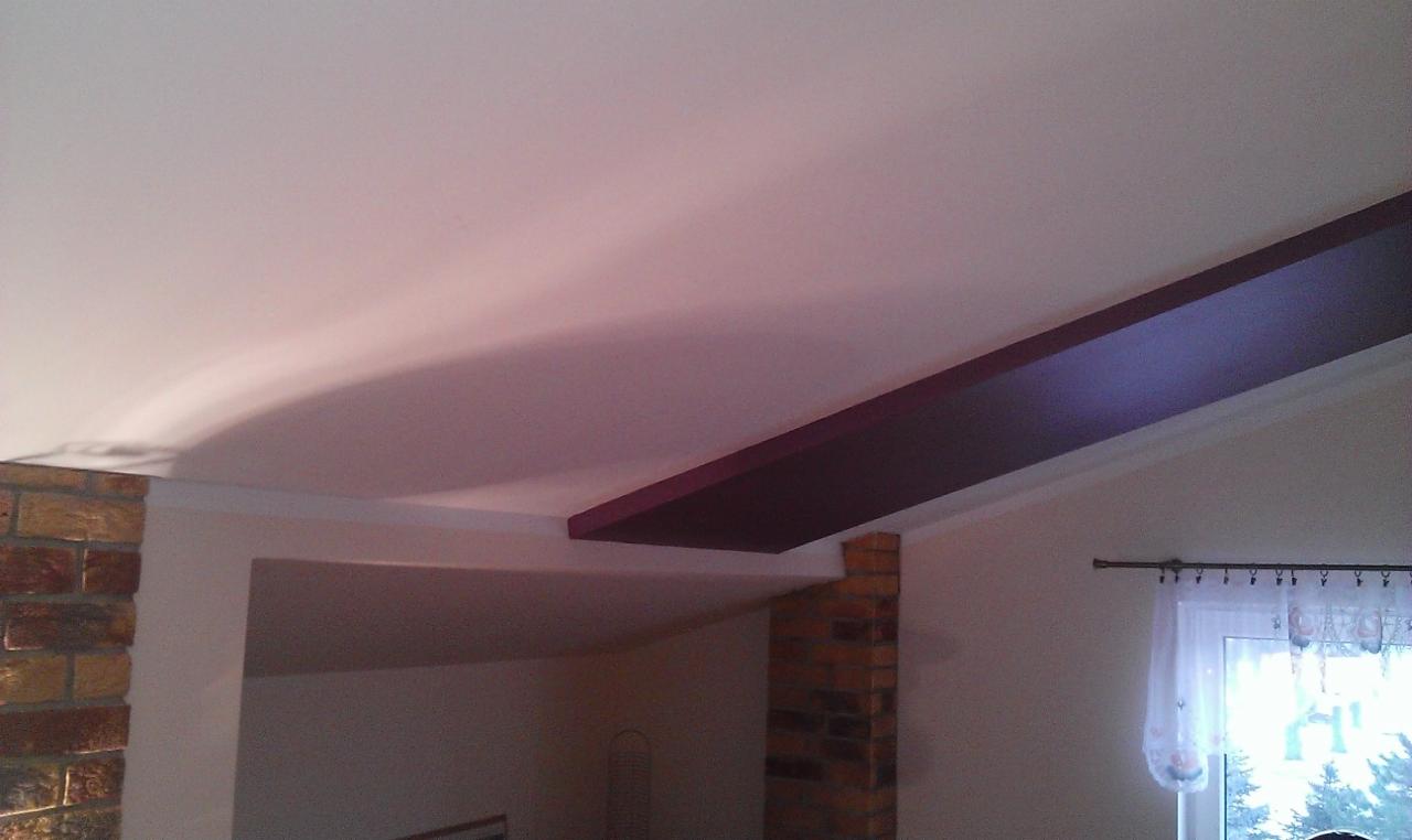 Jasny sufit z dekoracyjną, ciemną belką i ceglanym filarem. Widok na okno z firanką i fragment wnętrza. Detal wykończenia wnętrza po remoncie.