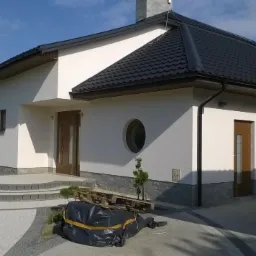 Elegancki, parterowy dom z białymi ścianami, ciemnym dachem i okrągłym oknem. Uwagę przykuwa starannie wykonany chodnik z kostki brukowej.