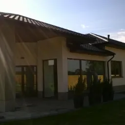 Elewacja domu jednorodzinnego w jasnym kolorze, z ciemną dachówką i dużymi oknami. Widoczne kolumny z elementami kamiennymi i zagospodarowany teren wokół budynku.