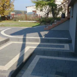 Ukończone schody i chodnik z kostki brukowej w odcieniach szarości, z dekoracyjnymi wstawkami z jasnego kruszywa. Widoczne balustrady i trawnik.