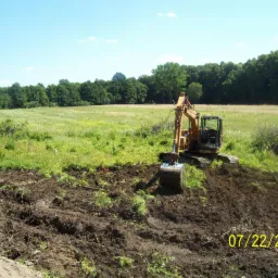 Żółta koparka na polu w trakcie robót ziemnych, widoczna data wykonania pracy. Z tyłu łąka i las.