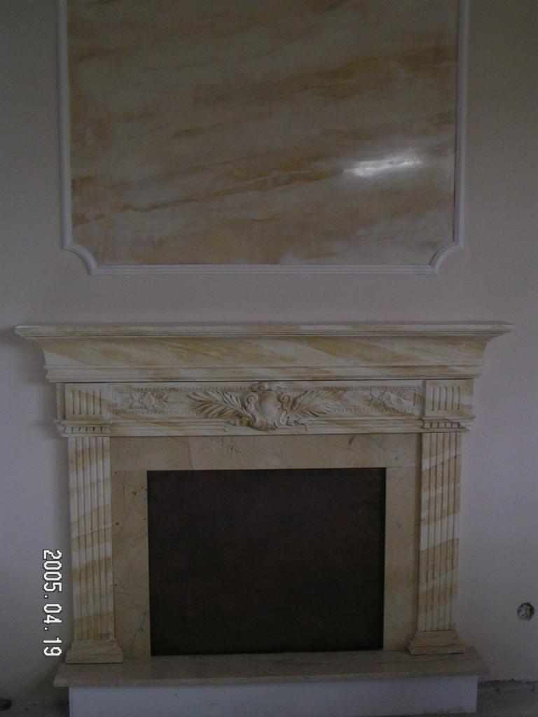 Klasycystyczny kominek z marmuru z dekoracyjnymi płaskorzeźbami i panel ścienny imitujący marmur, zamontowany w jasnym wnętrzu.