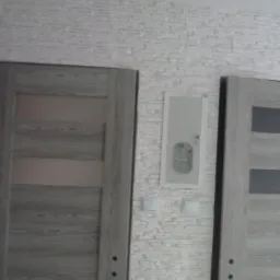 Dwie drewniane drzwi z przeszkleniami na tle ściany wykończonej dekoracyjną, białą cegłą, z widoczną skrzynką elektryczną i gniazdkami.