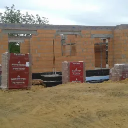 Budowa domu w trakcie wznoszenia ścian z czerwonej cegły Porotherm, z widocznymi otworami okiennymi i paletami cegieł na ziemi. Stan surowy otwarty.