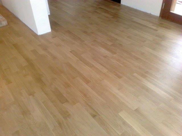 Jasna, lakierowana podłoga z parkietu ułożona we wzór cegiełki, widok z perspektywy podłogi, fragment ściany i drzwi w tle.