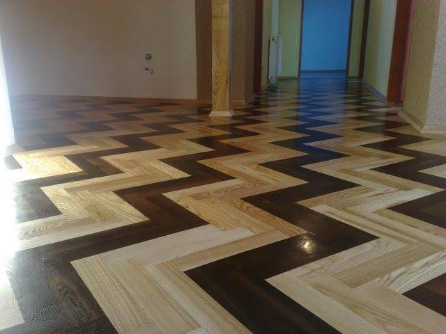 Parkiet ułożony w jodełkę z dwóch odcieni drewna: jasnego i ciemnego, tworzący zygzakowaty wzór na podłodze w korytarzu.