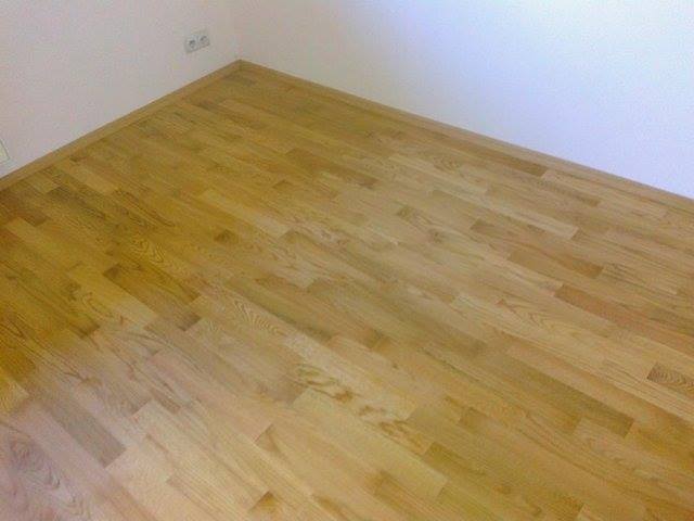 Jasny, lakierowany parkiet ułożony w jodełkę, widok z narożnika pomieszczenia z białymi ścianami i gniazdkiem elektrycznym.