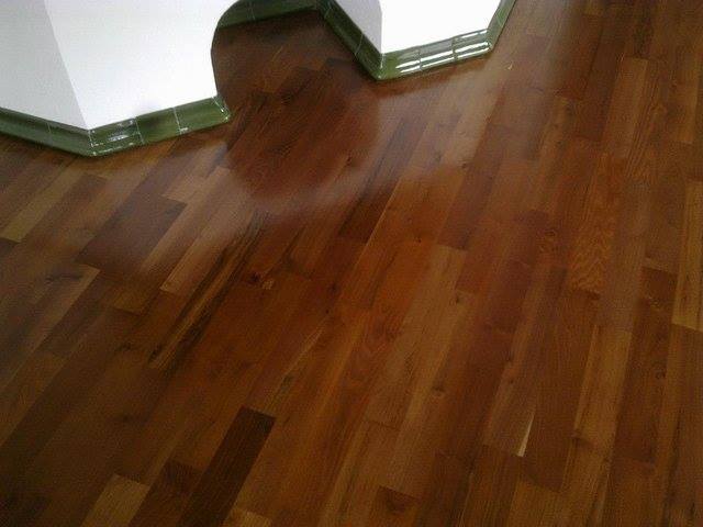 Błyszcząca, brązowa podłoga z parkietu ułożona pod kątem, z fragmentem białej ściany z zielonymi płytkami u dołu.