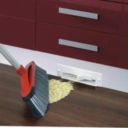 Szufelka automatyczna w zabudowie kuchennej, obok zmiotka z nagromadzonymi okruchami. Widoczna podłoga i dolna część szafek w kolorze burgundowym.