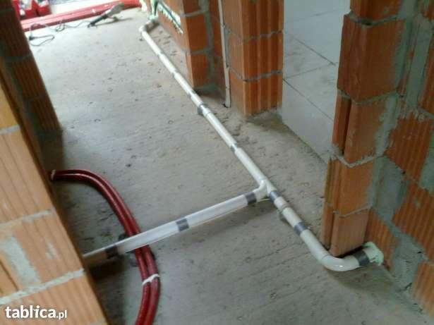 Białe rury instalacji centralnego odkurzacza, prowadzone w surowym stanie budynku, z widocznymi czerwonymi przewodami i fragmentem muru z cegły.