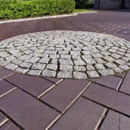 Okrągły wzór z kostki granitowej na tle ułożonej kostki brukowej w odcieniach czerwieni, otoczony zielenią.