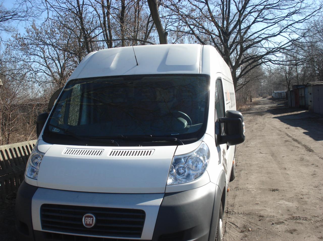 Biały bus marki Fiat na polnej drodze, widok z przodu na maskę i reflektory, w tle drzewa i blaszane garaże.