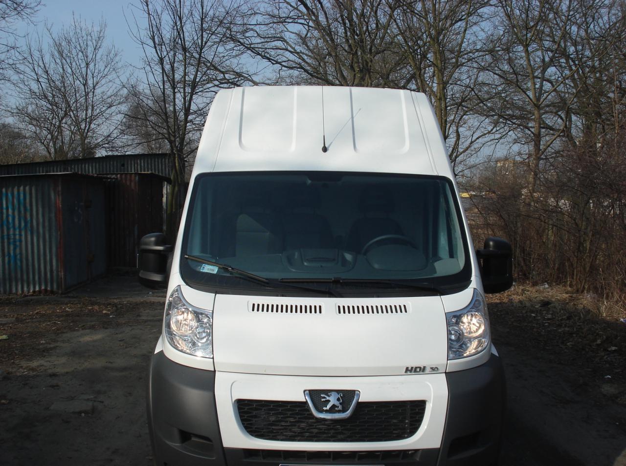 Biały bus dostawczy Peugeot Boxer HDI 30 sfotografowany z przodu na tle szarego blaszaka i drzew bez liści w pochmurny dzień.