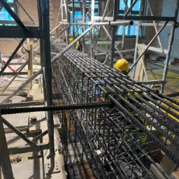 Zbrojenie pionowe z prętów stalowych, przygotowane do zalania betonem na budowie wewnątrz budynku, widoczne rusztowania i pracownik w żółtym kasku w tle.