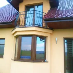 Beżowy dom z balkonem i dekoracyjną balustradą, widok z dołu. Okna z brązowymi ramami. Czerwona dachówka. Oświetlenie boczne.