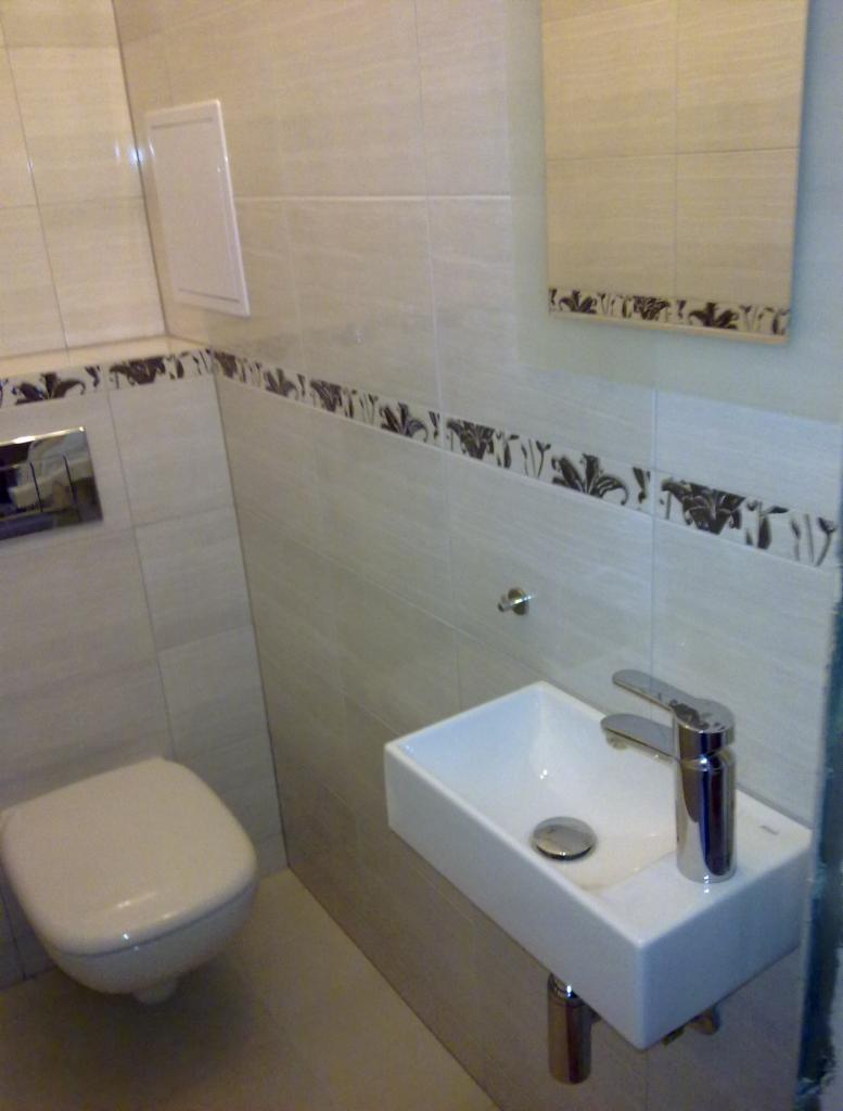 Wykończona łazienka z podwieszaną toaletą i małą, prostokątną umywalką z chromowaną baterią, wykończona beżowymi płytkami z dekoracyjnym pasem z motywem roślinnym.