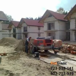 Pracownik obsługuje maszynę do wylewania posadzek na placu budowy z domami w tle. Widoczny piasek i materiały budowlane. Jasne światło dzienne.