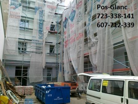 Budynek w trakcie prac remontowych, osłonięty siatką, widoczne rusztowania, kontener na odpady i samochód dostawczy. Prace elewacyjne w toku.