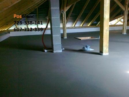 Szara, gładka wylewka na poddaszu z drewnianą konstrukcją dachu, widoczny otwór w podłodze i rura przy betonowym filarze. Wnętrze w trakcie wykończenia.