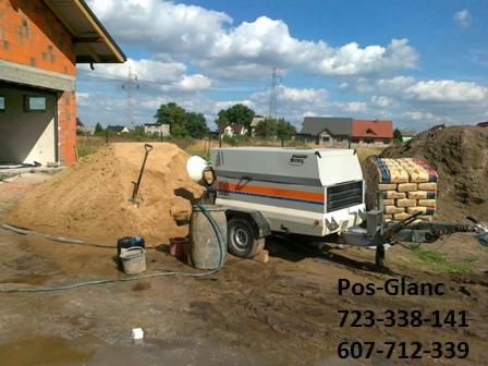 Sprzęt do wylewania posadzek na placu budowy: agregat z przyczepą, piasek, worki z materiałem. W tle budynek w trakcie budowy i błękitne niebo.