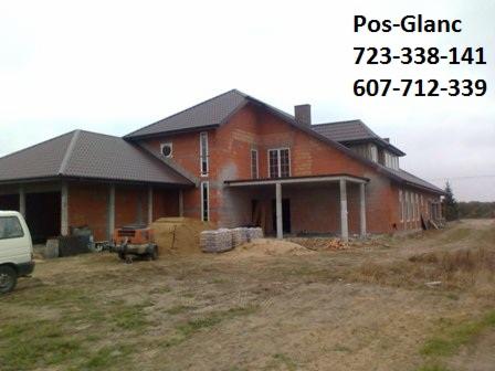 Surowy stan budynku z czerwonej cegły z brązowym dachem, widoczne materiały budowlane i sprzęt. Na górze numer telefonu do firmy Pos-Glanc.