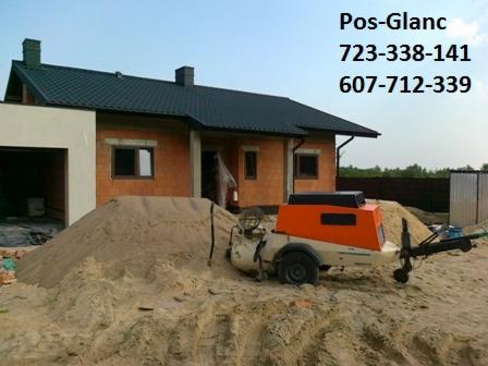 Pompa do wylewek stoi obok sterty piasku na tle domu w trakcie budowy. Widoczne numery kontaktowe firmy Pos-Glanc.