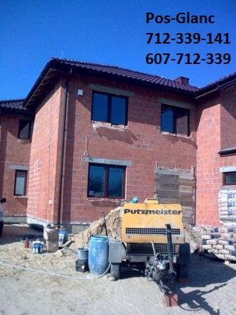 Żółta maszyna Putzmeister na placu budowy, obok budynku z czerwonej cegły i worków cementu. Widoczne okna i dach. Błękitne niebo w tle.