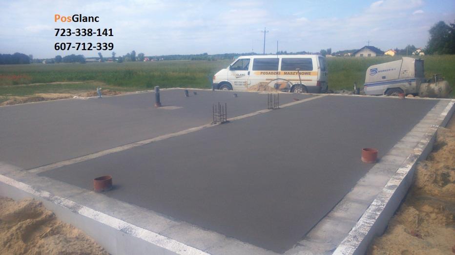 Świeżo wylana, gładka posadzka na fundamencie z wystającymi rurami i zbrojeniami. W tle samochód z napisem 'Posadzki Maszynowe' i maszyna do wylewania.