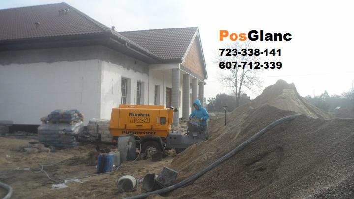 Prace wylewkowe na budowie domu jednorodzinnego. Żółta maszyna Mixokret i stosy piasku. Pracownik w niebieskiej bluzie obsługuje urządzenie. Widoczne worki z materiałami.