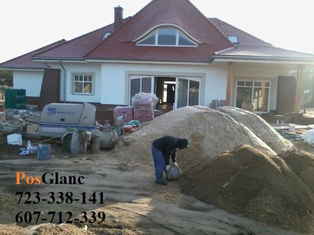 Prace przygotowawcze do wylewki: pracownik nabiera zaprawę obok maszyny do posadzek na tle domu z czerwoną dachówką. Widoczne materiały budowlane.