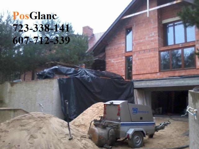 Betoniarka przy stercie piasku na budowie domu z czerwonej cegły. Widoczne narzędzia i materiały budowlane. Częściowo zadaszony teren budowy.