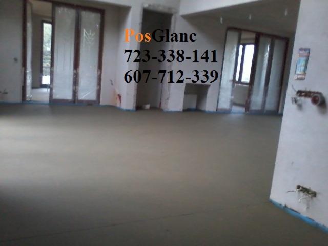 Wnętrze z wylaną, gładką posadzką w odcieniach szarości. Widoczne przygotowane ściany i drzwi przesuwne. Ujęcie z niskiego kąta podkreśla równość powierzchni.