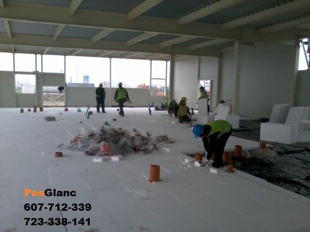 Montaż izolacji podłogowej z rurami i styropianem w hali przemysłowej. Pracownicy w kamizelkach odblaskowych układają elementy na białej powierzchni.