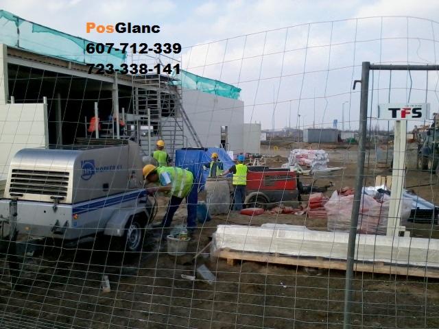 Prace na budowie: ekipa w żółtych kamizelkach obsługuje maszynę do wylewek, widoczne materiały budowlane i ogrodzenie.