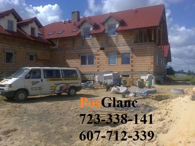 Budynek w trakcie wylewania posadzki maszynowej, widoczny bus z logo firmy, betoniarka i pracownik. Czerwony dach i ceglana elewacja w surowym stanie.