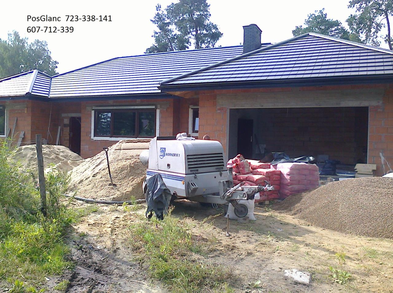 Budowa domu w trakcie: agregat do wylewek stoi obok sterty piasku i worków z zaprawą, w tle surowy budynek z cegły z widoczną konstrukcją dachu.