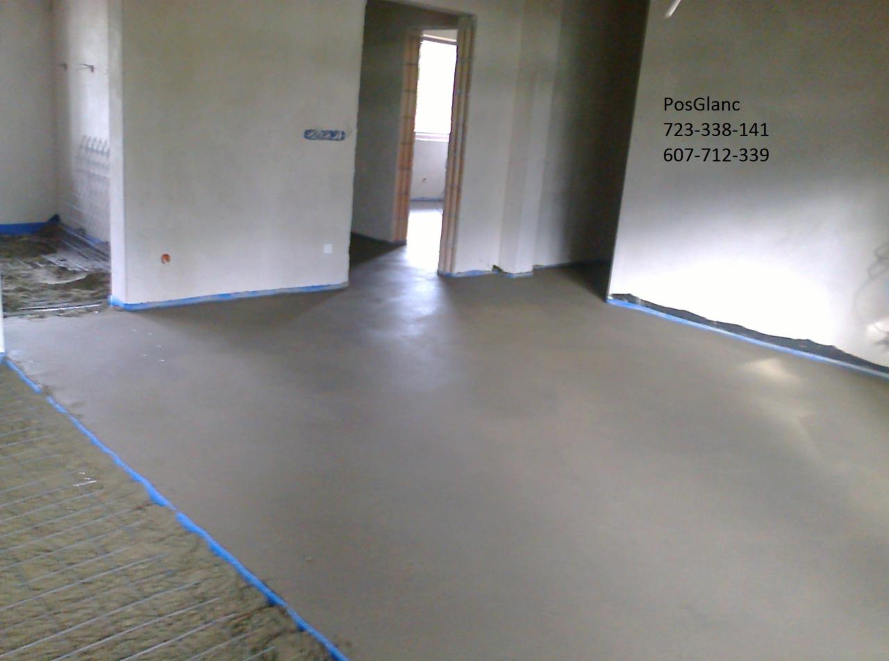 Świeżo wylana, gładka posadzka jastrychowa w jasnym pomieszczeniu z widoczną siatką zbrojeniową przy ścianie. Niebieska taśma maskująca zabezpiecza ściany przed zabrudzeniem.