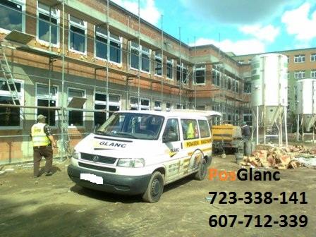 Praca na budowie: biały bus firmy Glanc z pracownikiem w kamizelce odblaskowej na tle budynku w trakcie remontu i silosów. Widoczne rusztowania i materiały budowlane.