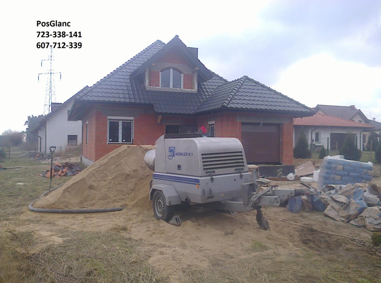 Agregat do wylewek stoi na placu budowy przed domem z czerwoną cegłą. W tle widoczny piasek i worki z materiałem budowlanym. Dzień pochmurny.