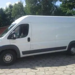 Biały bus dostawczy marki Citroen Jumper, zaparkowany na brukowanej nawierzchni, z zielonymi drzewami w tle. Widok z boku, lekko z przodu.