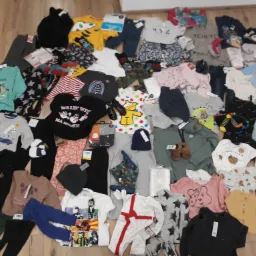 Stos ubrań dziecięcych z metkami ułożony na drewnianej podłodze, obejmujący różnorodne wzory i kolory, od bluzek z nadrukami po spodnie i piżamy, z widocznymi detalami jak małe buciki i czapki.