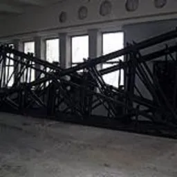 Szkielet metalowej konstrukcji w hali, widoczne spawane elementy, w tle okna z białymi ramami, szara betonowa podłoga.